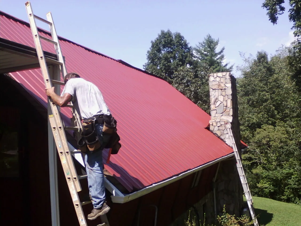 Roofer on a ladder beside a red metal roof and stone chimney — Soffit & Fascia Repair by Roofing Brothers in Ayr